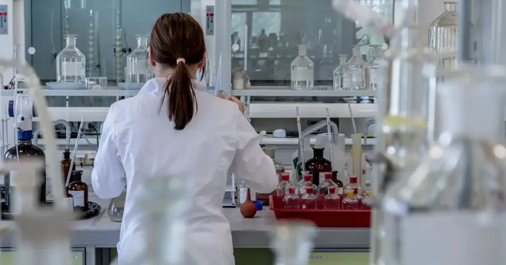 Investigadora con bata blanca, de espaldas, trabajando en un laboratorio con frascos y material de vidrio